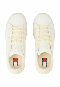 Baskets blanches avec une tige synthétique lisse, des lacets jaunes, une boîte à orteils texturée, et une étiquette de logo sur la semelle intérieure affichant "TOMMY JEANS."