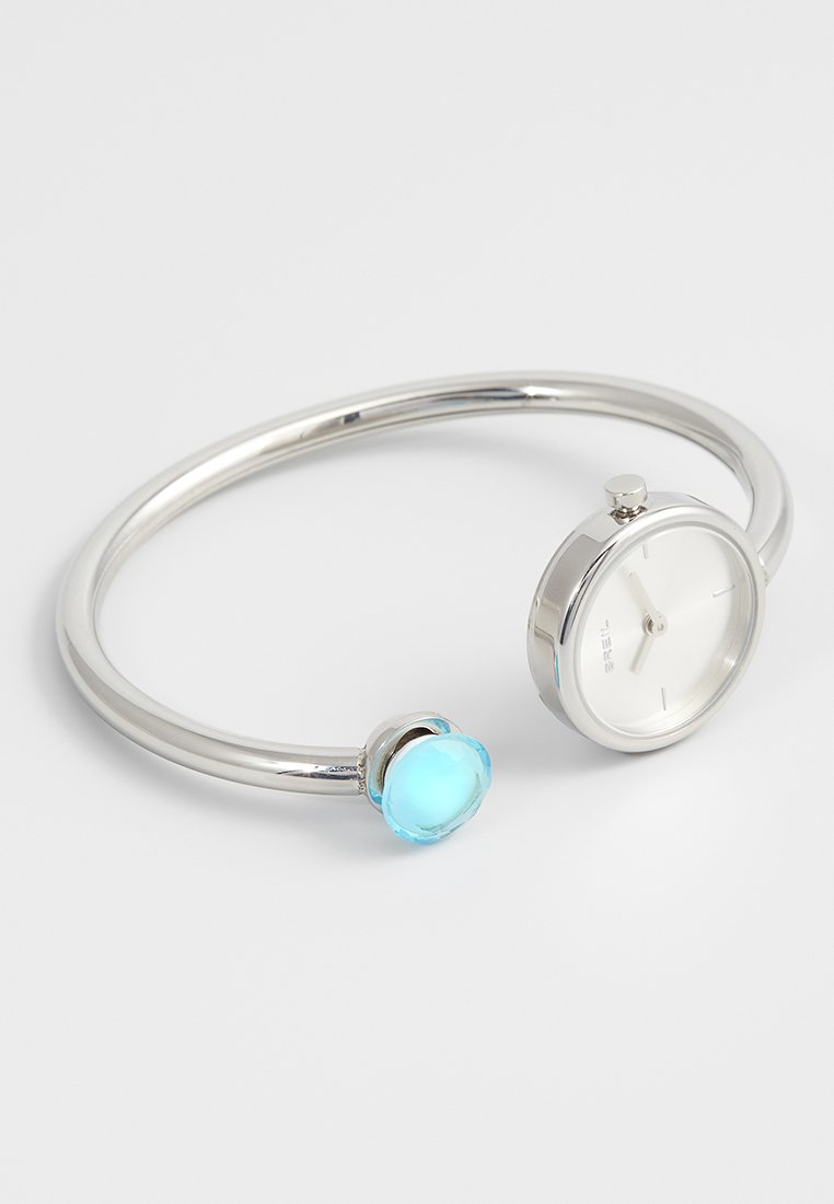 Zilveren bangle-horloge met witte wijzerplaat, minimalistisch design, accenten van een blauwe edelsteen. Glad, rond vorm. Voorzien van een eenvoudige metalen sluiting.