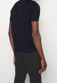 Kortärmad marinblå stickad tröja med ribbad nederkant och manschetter, med detaljer på axlarna. Kombineras med mörka byxor. Texturerad tyg.