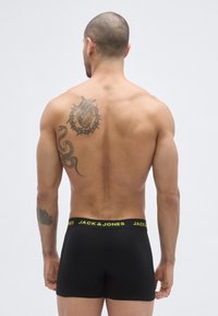 Boxer noir avec une ceinture jaune marquée, offrant une coupe ajustée. Peau visible dans le dos du modèle, montrant des tatouages.