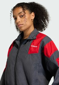 Siva lagana jakna s crvenim detaljima, s visokim ovratom, zatvaranjem na zip, te Adidas logom na prsima. Glatka tekstura.