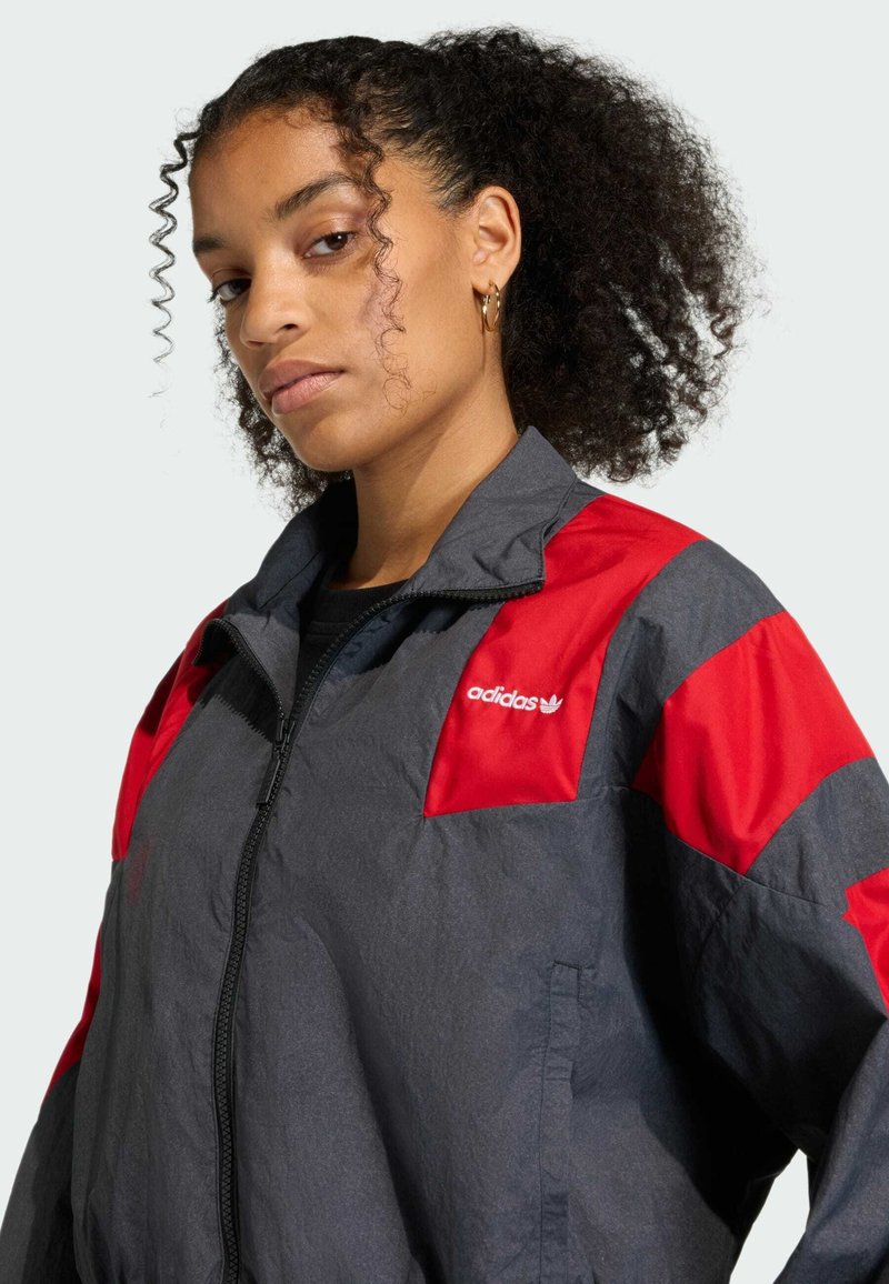 Graue, leichte Jacke mit roten Akzenten, hoher Kragen, Reißverschluss und dem Adidas-Logo auf der Brust. Glatte Textur.