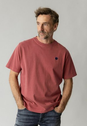 Mand med let skæg iført en falmet rød T-shirt med korte ærmer og blå jeans med hænderne i lommerne, der kigger til siden mod en ensfarvet baggrund.