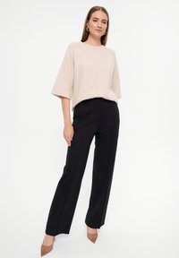 Hellbeige, kurzärmeliger Strickpullover in entspannter Passform, kombiniert mit hochgeschnittenen, maßgeschneiderten schwarzen Hosen und nackten Stilettos.