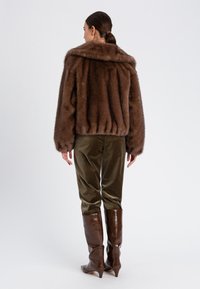 Giacca marrone in pelliccia sintetica con ampio colletto, orlo a costine e maniche voluminose, abbinata a pantaloni in velluto verde oliva e stivali marroni al ginocchio.