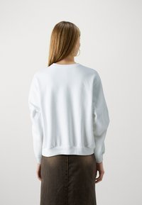 Λευκή μπλούζα με χαλαρή εφαρμογή, ριμπ μανσέτες και ζώνη. Λείος υφή, στρογγυλή λαιμόκοψη και μινιμαλιστικός σχεδιασμός σε ανοιχτό φόντο.