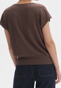 Blusa marrón texturizada de manga corta, con un patrón de cuadrícula, ajuste holgado y dobladillo acanalado. Combinada con denim oscuro.