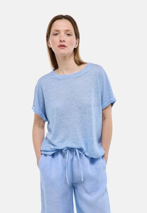 Femme portant un haut ample à manches courtes bleu clair et un pantalon à cordon assorti, debout avec les mains dans les poches devant un fond blanc.