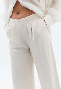 Beige veckade byxor i lätt material, med sidofickor och en avslappnad passform, kombinerade med en något genomskinlig, ljusblommig topp.