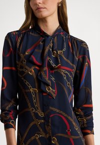 Γυναίκα με μπλε μπλούζα με δεμένο φιόγκο στον λαιμό και σχέδιο από κόκκινες, καφέ και χρυσές αλυσίδες και ζώνες, με τα μανίκια σηκωμένα μέχρι τους πήχεις.