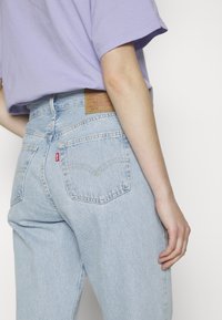 Personne portant un jean Levi's taille haute bleu clair et une chemise lavande à manches courtes, vue de mi-dos à mi-cuisse.