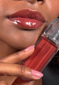 Gloss à lèvres 3D Hydra Xtreme dans un tube rectangulaire transparent avec un capuchon argenté. Teinte rouge profond, brillante, appliquée sur des lèvres pulpeuses avec une subtile brillance.