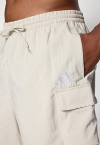 Beige cargoshorts med elastisk midja, dragsko, sidoficka och vit broderad logotyp på vänster lår. Slät tygtextur.