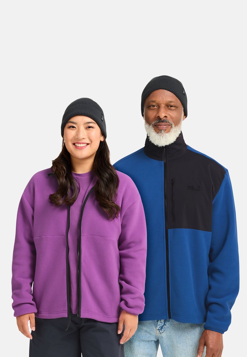 Two individuals stand side by side, wearing fleece jackets. The woman has a purple jacket, while the man wears a blue and black jacket. Both have dark knit beanies.