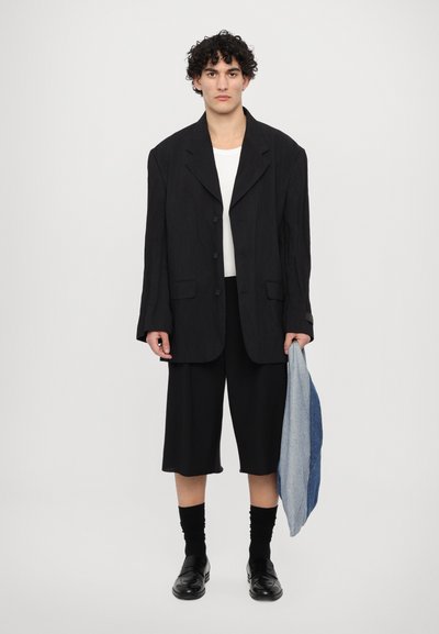 Černá oversized sako s velkými klopami, doplněná černými šortkami do délky kolen. Model drží džínovou tašku a má na sobě černé ponožky a mokasíny.