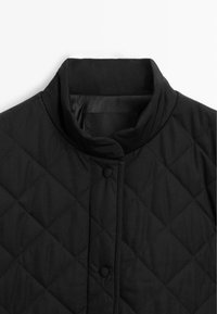 Colletto di giacca nera trapuntata con bottoni a pressione, caratterizzato da una texture di tessuto liscia e un sottile motivo a diamante. Design minimalista.