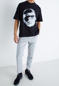 Black short-sleeve t-shirt with a blurred face graphic, paired with light gray slim-fit trousers and black sneakers with textured detailing.