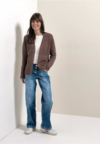 Cardigan côtelé marron porté sur un haut blanc, associé à un jean large bleu clair et des baskets blanches. Design simple avec une coupe décontractée.