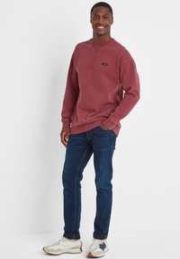 Smiling man wearing a maroon sweatshirt, dark blue jeans, and white sneakers, standing with one hand in pocket against white background.