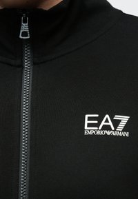 Czarny strój zapinany na zamek z białym logo EA7 Emporio Armani na piersi oraz suwakiem z wytłoczonym logo EA7.