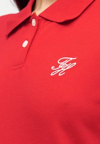 Κόκκινο πόλο πουκάμισο από υφασμάτινη υφή, με ριμπ γιακά και δύο κουμπιά. Ένα κεντημένο λογότυπο εμφανίζεται στο αριστερό μέρος του στήθους.