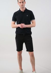 Chemise polo noire avec des accents rayés blancs sur les manches, associée à un short noir. Le mannequin porte des chaussures slip-on grises claires.