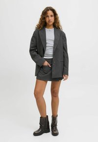 Mladiž ženska s kodrasto pričesko, oblečena v sivo blazer, ujemajočo se krilo, svetlo sivo majico in temno rjave gležnjarje, ki stoji ob belem ozadju.