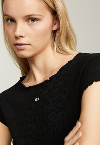 Top negro texturizado con borde ondulado y mangas cortas. Presenta un pequeño logo bordado en el pecho. Primer plano de la prenda y el modelo.