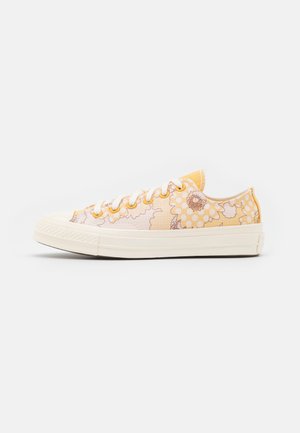 Baskets basses en toile avec motif floral jaune et beige, lacets blancs et semelle en caoutchouc écru sur fond blanc.