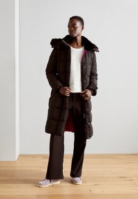 Abrigo de plumas negro largo con capucha forrada de piel, con un interior rosa, combinado con un suéter blanco y pantalones oscuros. Fondo de suelo de madera.