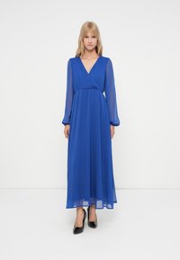 Mulher loira em pé, vestindo um vestido azul royal de comprimento total e mangas longas com decote em V, e sapatos de salto alto pretos, em um fundo branco.