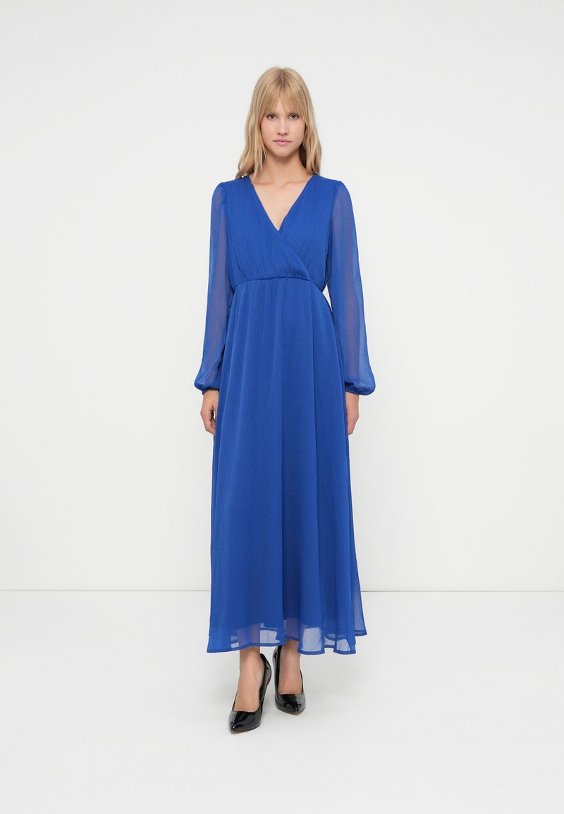 Mulher loira em pé, vestindo um vestido azul royal de comprimento total e mangas longas com decote em V, e sapatos de salto alto pretos, em um fundo branco.