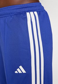 Mėlynos sportinės šortai, pagaminti iš lygaus poliesterio, su trimis vertikaliomis baltomis juostelėmis ir mažu baltu logotipu kairėje pusėje.