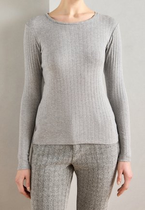 Frau trägt eng anliegendes graues, geripptes Langarmshirt und graue Hose mit Fischgrätmuster, steht vor einem schlichten Wandhintergrund.