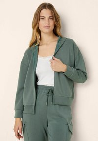 Felpa con cappuccio e zip in verde tenue, realizzata in tessuto morbido. Presenta una vestibilità comoda, tasche frontali e è abbinata a una maglietta bianca con rifinitura in pizzo.