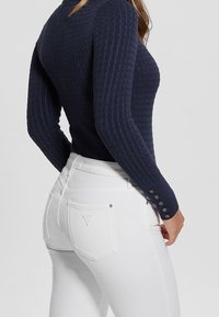 Marineblaue Rippstrickpullover mit strukturiertem Muster und Knopfakzenten an den Ärmeln, kombiniert mit hochtaillierten, weißen Skinny-Jeans mit Logodetail.