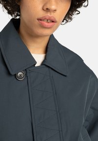 Close-up of a person wearing a dark gray collared coat with a button near the neck and zigzag stitching on the front flap.