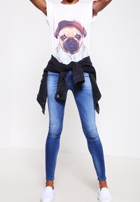 Camiseta blanca con un gráfico de un pug, chaqueta de mezclilla negra atada a la cintura, jeans ajustados azules y zapatos slip-on blancos.