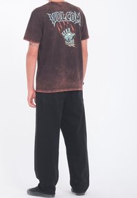 Henkilö, joka on pukeutunut haalistuneeseen mustaan Volcom T-paitaan, jossa on sininen käsi ja punaiset kynnenjäljet grafiikka selässä, yhdistettynä olohuoneen mustiin housuihin ja tennareihin.