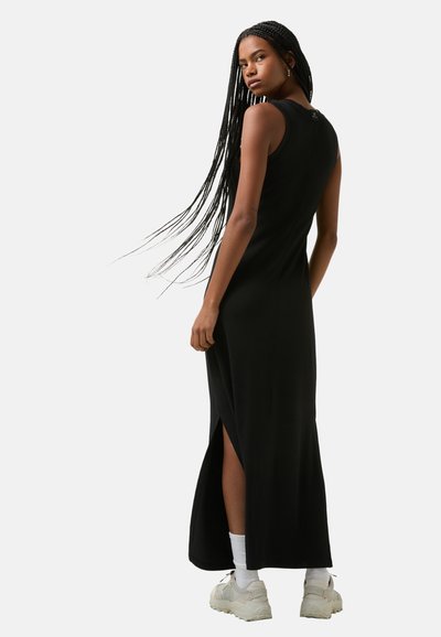 Zwarte mouwloze maxi-jurk met zijslits, geribbelde textuur, aansluitend ontwerp en een open rug. Draagt met witte sokken en chunky sneakers.