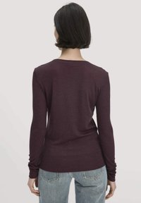 Frau mit kurzem, dunklem Haar, die ein langärmliges lila Oberteil und hellblaue Jeans trägt, steht mit dem Rücken zu einem schlichten Hintergrund.