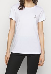 Bílý bavlněný tričko s krátkým rukávem, kulatým výstřihem a malým černým vzorem listu na hrudi. V kombinaci s černými spodními díly.