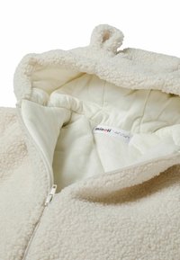 Veste en polaire crème avec un extérieur texturé et doux, une capuche et une doublure lisse. Dispose d'une fermeture éclair à l'avant et d'une étiquette intérieure.
