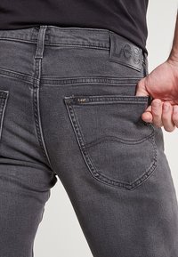 Šedé džíny z denimu s přiléhavým střihem, které mají viditelnou zadní kapsu a texturovanou koženou nášivku na pásku.