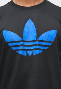 Melna kokvilnas T-krekls ar lielu zilu Adidas trifīsādas logotipa grafiku ar trīs svītrām apakšā, kas attēlo ģeometrisku tekstūru.