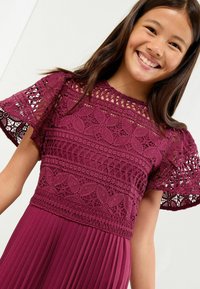 Rochie burgundy cu detalii intricate din dantelă pe corset și mâneci flotante, având o fustă plisată pentru un plus de textură și mișcare.
