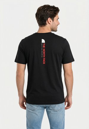 Hombre de espaldas con una camiseta negra de The North Face con logo vertical rojo y blanco en la parte trasera y vaqueros azul claro.