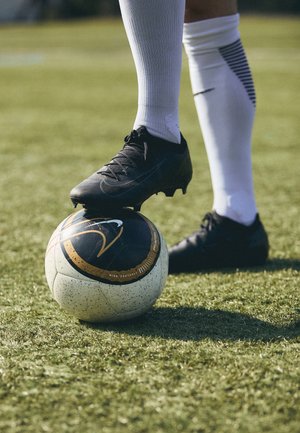 Schwarzer Stollenschuh für Fußball auf einem weißen Sockel tragenden Fuß, der auf einem schwarz-weißen Nike-Fußball auf einem Grasfeld ruht.