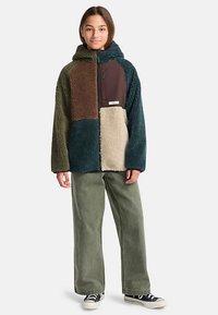 Sweat à capuche en polaire avec un design patchwork en marron, vert et beige. Comprend une poche zippée et une coupe décontractée. Associé à un pantalon large de couleur olive.