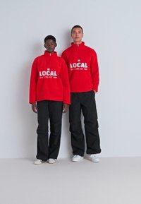 Pullover in pile rosso con colletto alto e testo bianco, abbinati a pantaloni neri larghi. Gli outfit dei modelli presentano un design casual.